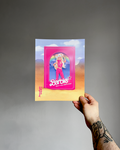 Hand holding an art print with a Barbie design against a gray background