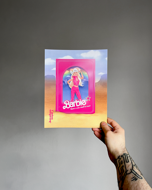 Hand holding an art print with a Barbie design against a gray background