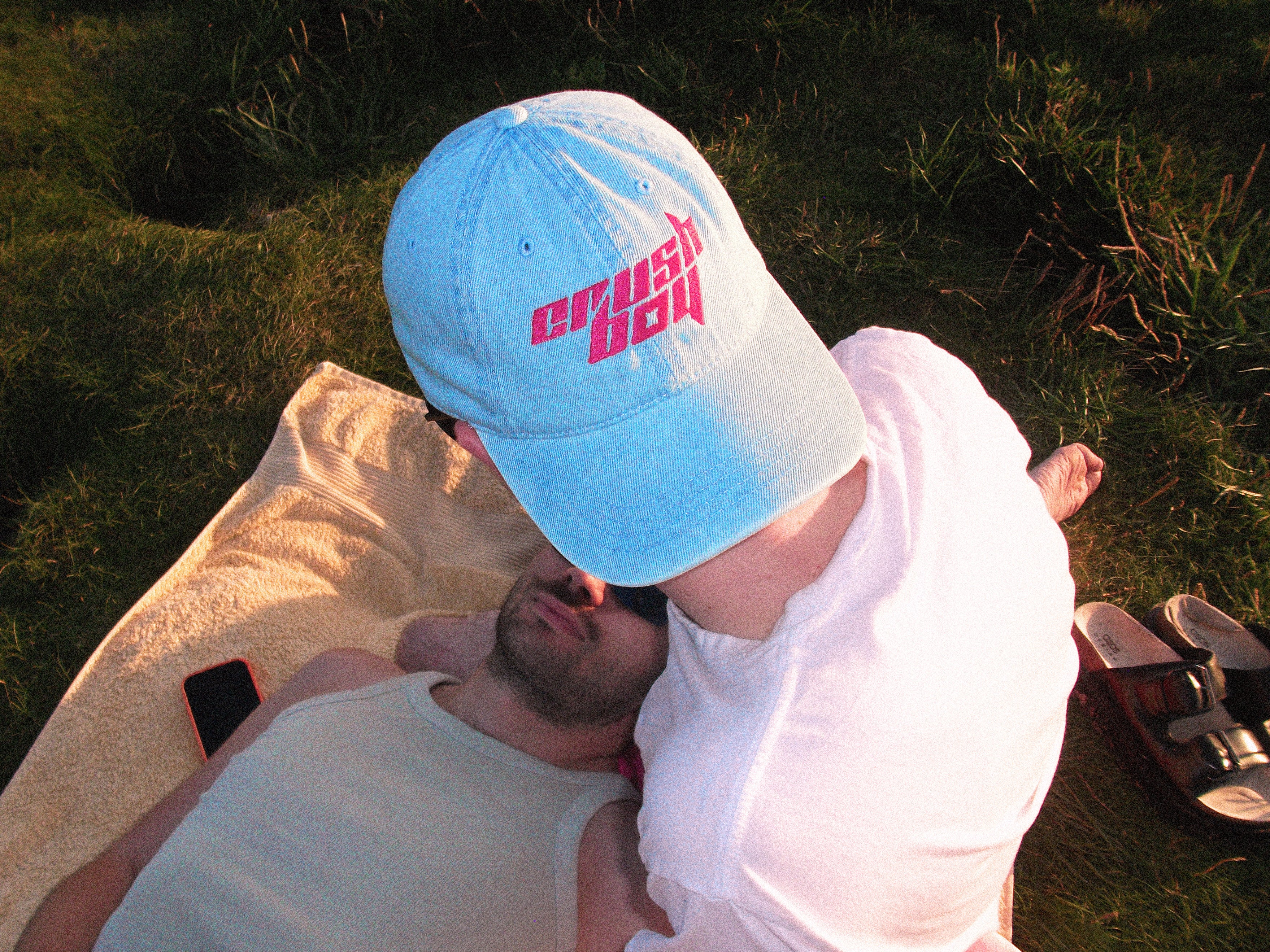 A man wearing a light blue baseball cap with pink 'CRUSHBOY' text leans in close to another man lying on a blanket outdoors on grass.