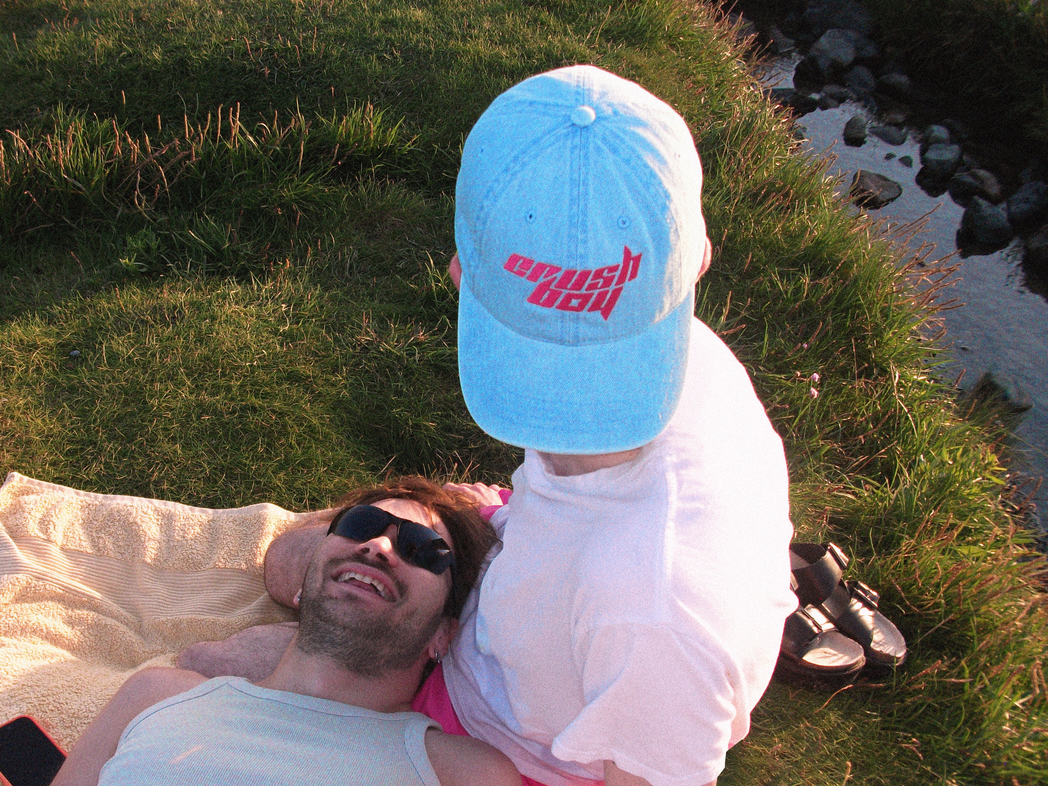 A man wearing a light blue denim baseball cap with pink stylized text that reads 'CRUSHBOY' on it and a white t-shirt sits on a grassy lawn next to a body of water, leaning on another man lying on a blanket.