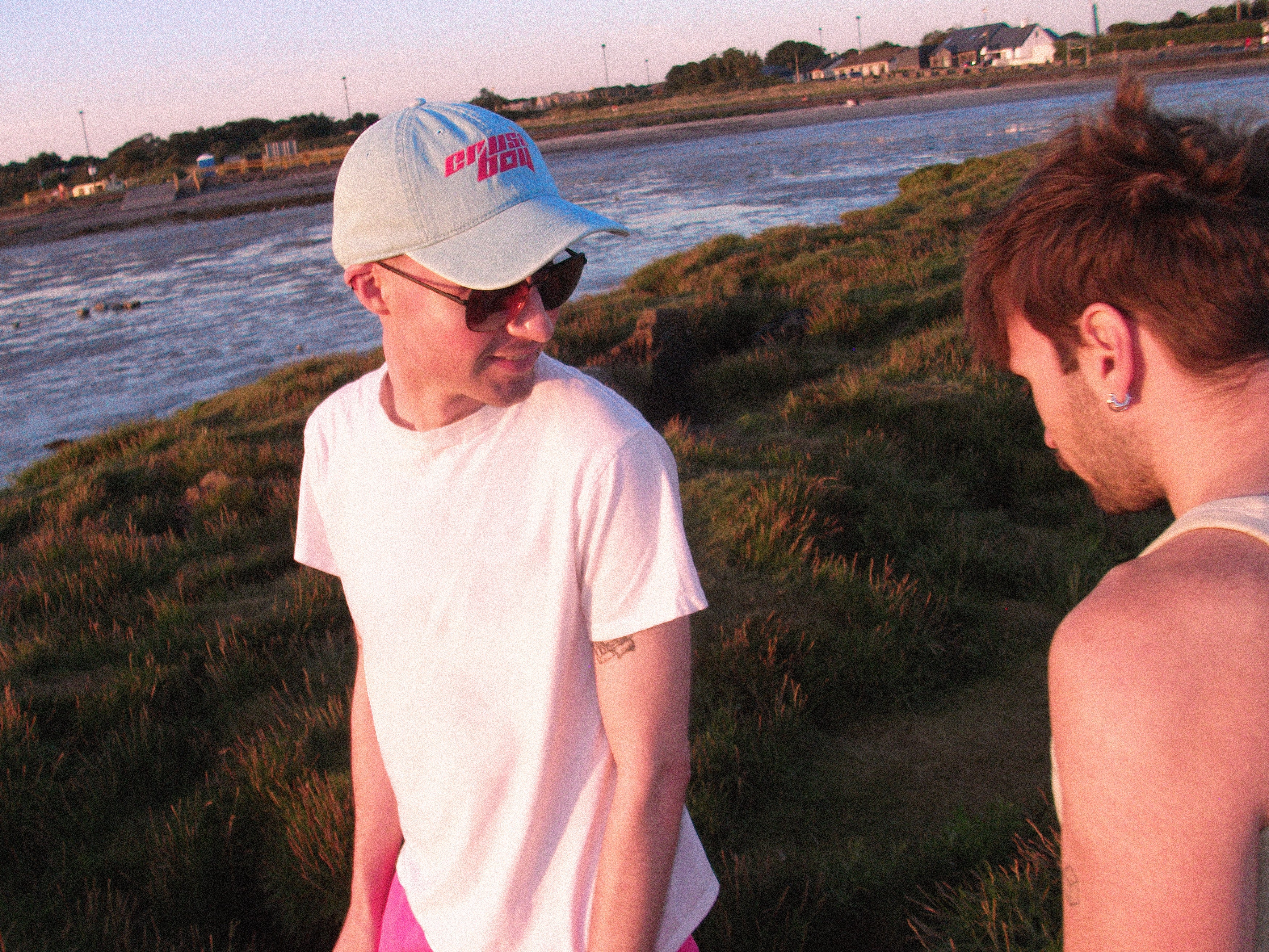Two men standing outdoors near a beach at sunset. One man is wearing a white t-shirt, pink shorts, sunglasses, and a light blue denim baseball cap with pink text that reads 'CRUSHBOY'.