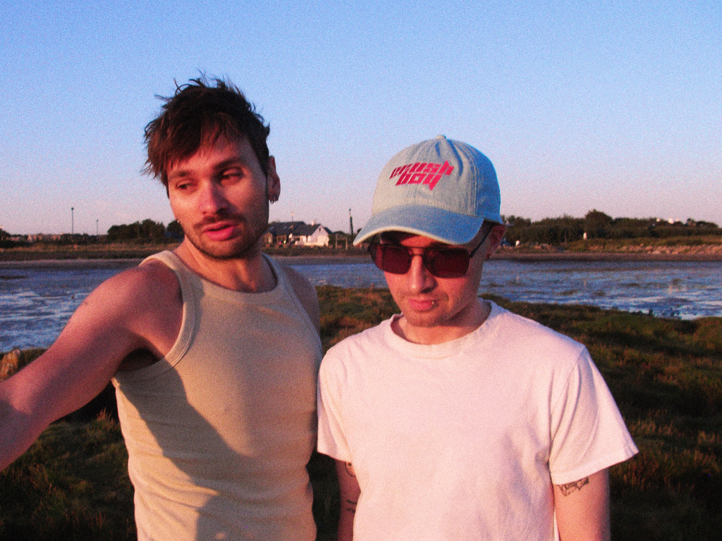 Two men standing outdoors near a beach at sunset, one wearing a beige tank top and the other wearing a white t-shirt, sunglasses, and a light blue denim baseball cap with 'CRUSHBOY' on it in pink text.