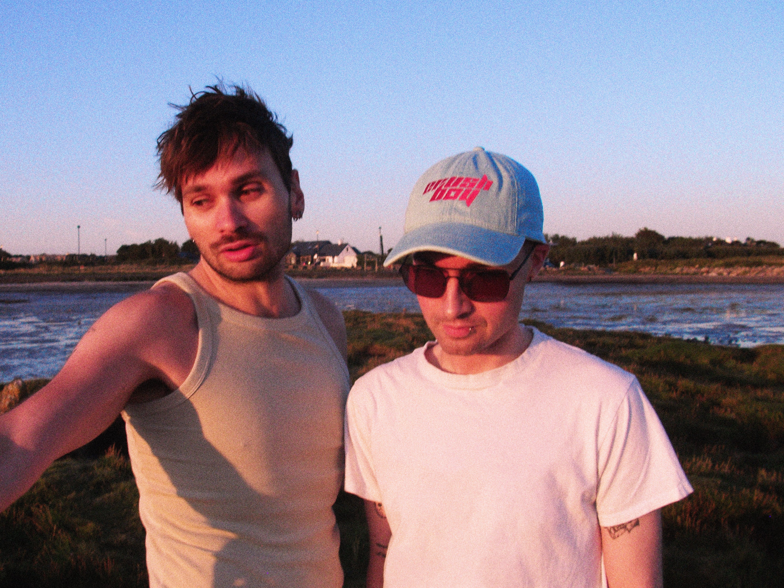 Two men standing outdoors near a beach at sunset, one wearing a beige tank top and the other wearing a white t-shirt, sunglasses, and a light blue denim baseball cap with 'CRUSHBOY' on it in pink text.