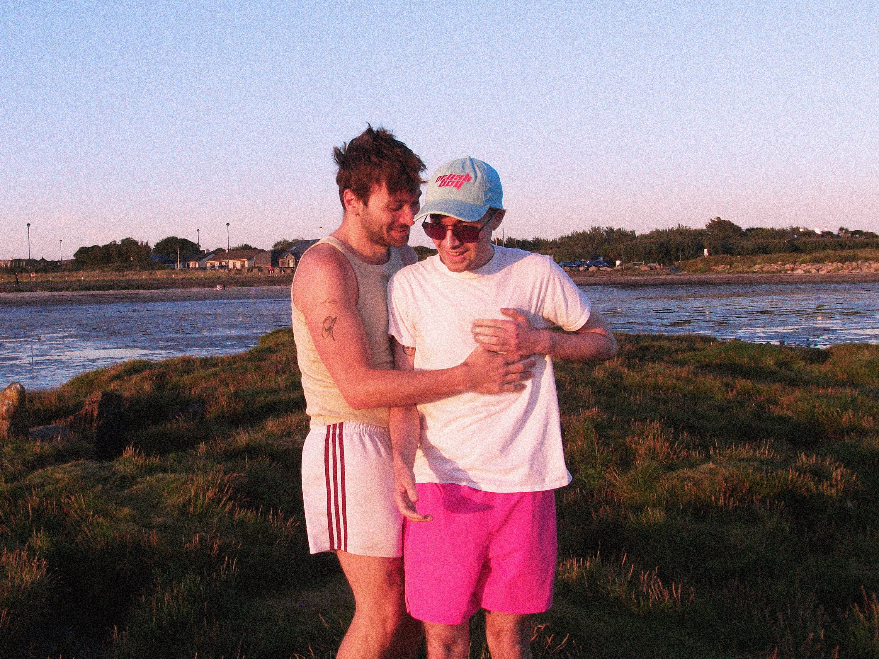 Two men embrace outdoors by a beach at sunset, wearing casual swimwear. One wears a beige tank top, white shorts with red stripes, and white sneakers.