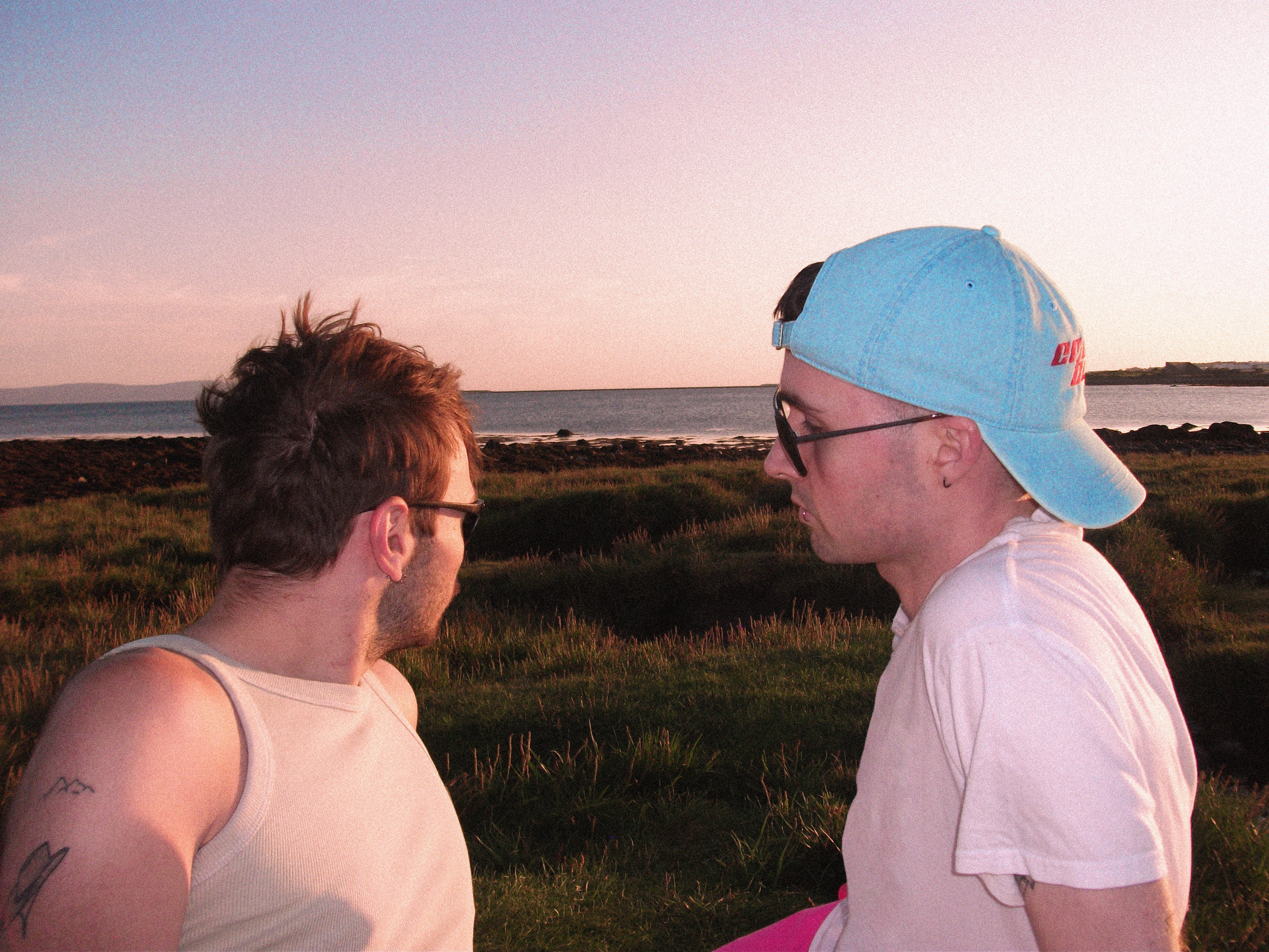 Two men sitting outdoors by the beach at sunset, facing each other. One man wears a light blue baseball cap, black sunglasses, a white t-shirt, and pink shorts.