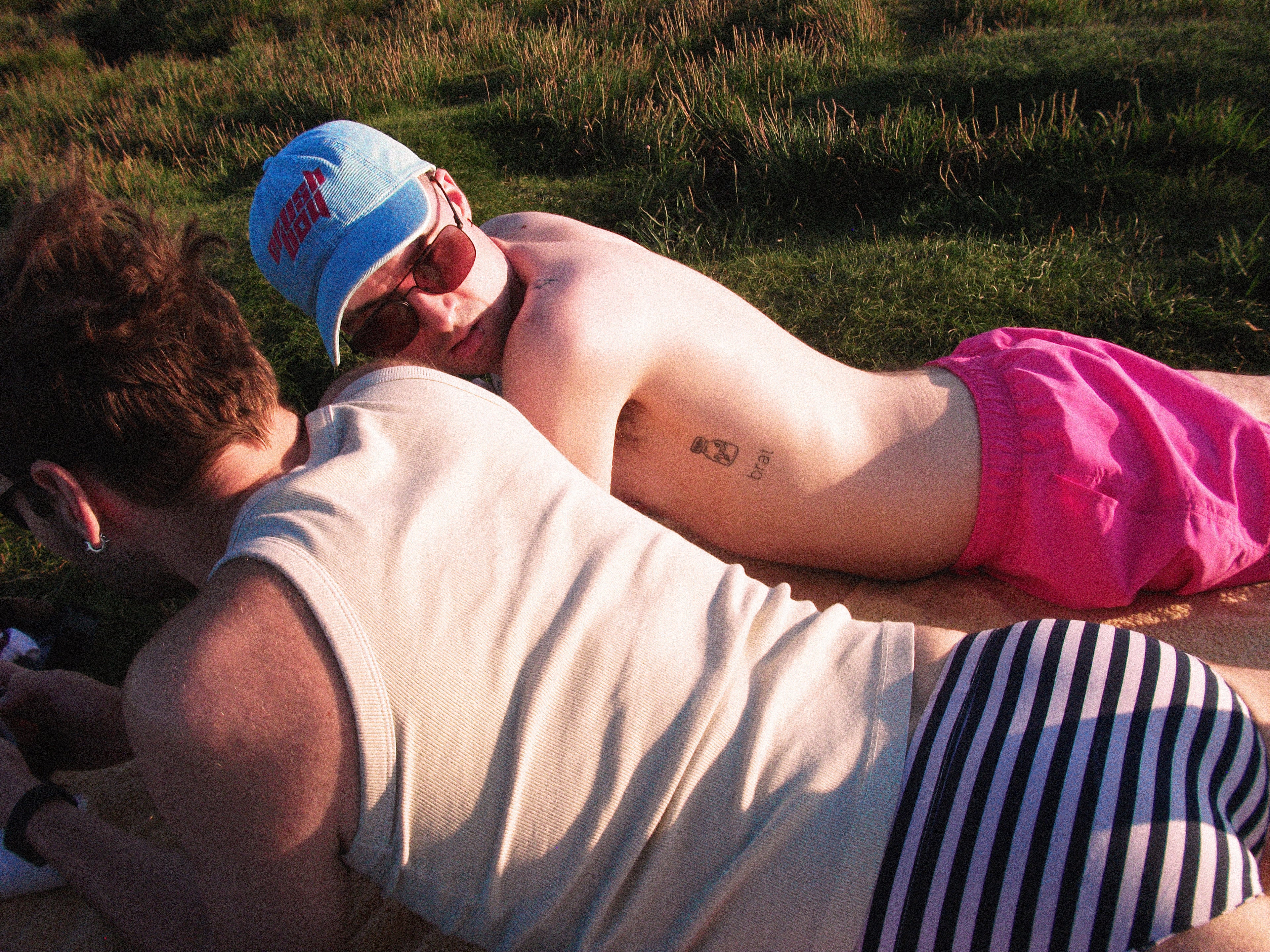 Two men lying on a blanket outdoors on a grassy lawn. One man is wearing a light blue denim baseball cap with pink lettering that reads 'CRUSHBOY', pink swim trunks, and sunglasses, while the other wears a beige tank top and black-and-white striped swim briefs.