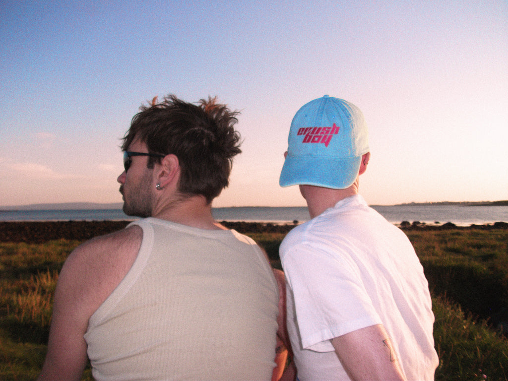 Two men sitting by the beach at sunset, with one wearing a light blue baseball cap with pink text that reads 'CRUSHBOY' and the other wearing a beige tank top.