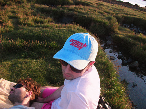 A man wearing a light blue denim baseball cap with a pink 'CRUSHBOY' logo and sunglasses sits outdoors on a grassy hill next to the beach, with another man lying down on his lap.