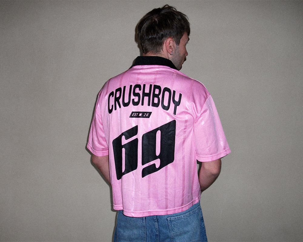 A man viewed from behind wearing a pink short-sleeve polo shirt with 'CRUSHBOY EST. 26' and the number '69' printed in black on the back, paired with blue jeans against a plain gray wall.