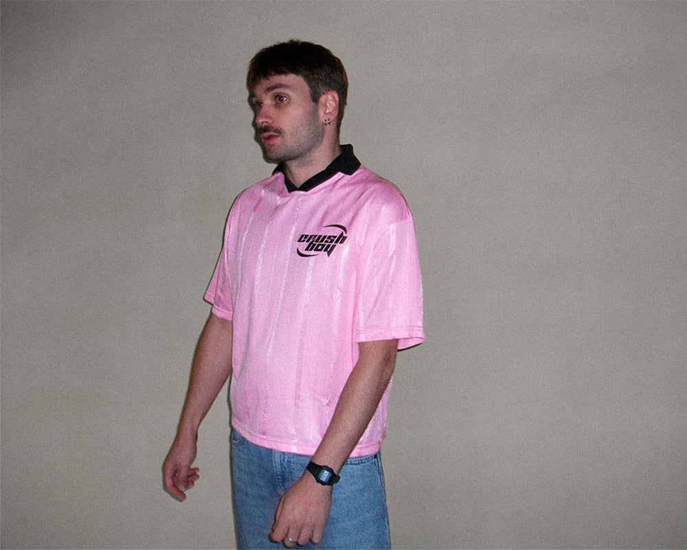 A man wearing a light pink short-sleeve polo shirt with a black collar and a black 'Porsche' logo on the chest, paired with blue jeans, standing against a plain gray wall.