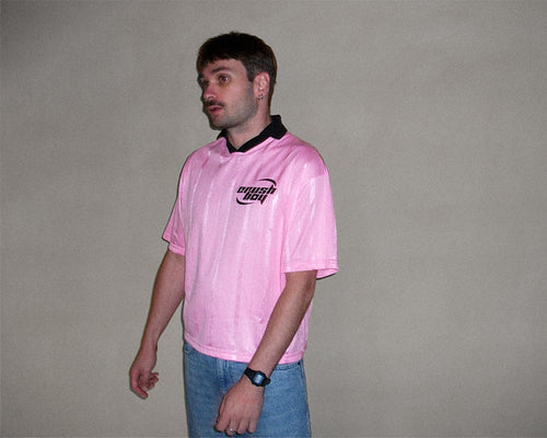 A man wearing a light pink short-sleeve polo shirt with a black collar and a black 'Porsche' logo on the chest, paired with blue jeans, standing against a plain gray wall.