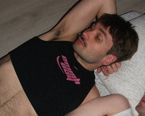 A man lying down wearing a black sleeveless tank top with a pink 'crushboy' graphic on the chest, photographed in dim indoor lighting.