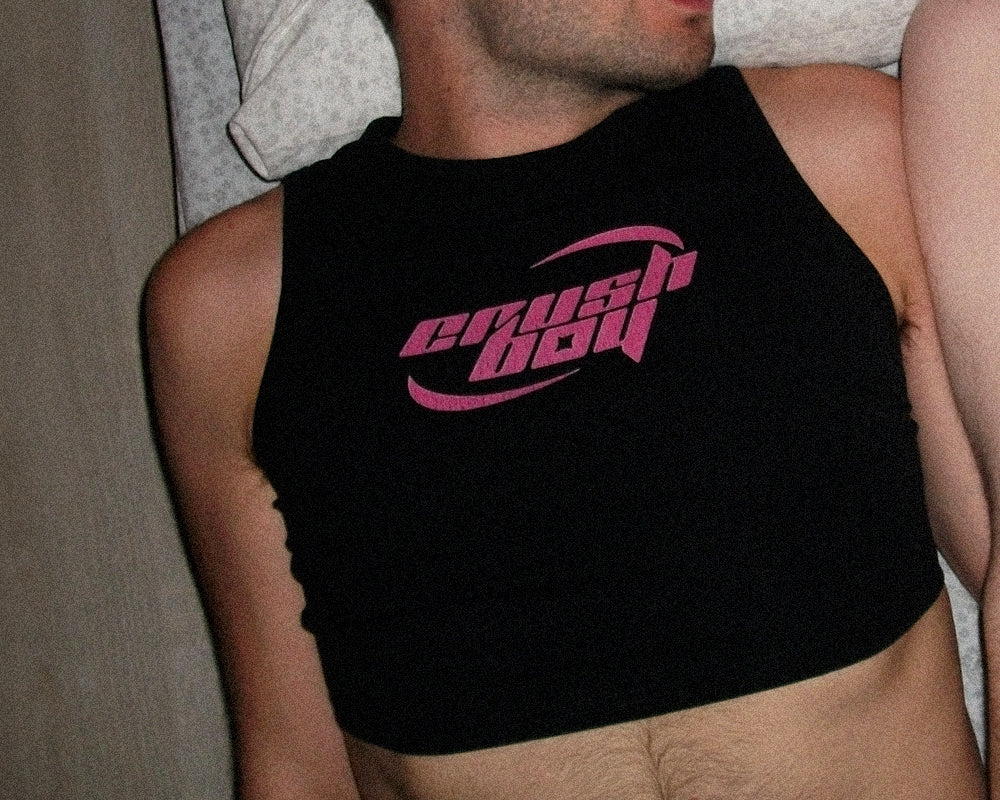 A close-up of a man wearing a black sleeveless crop top with a pink 'crushboy' graphic on the chest, photographed indoors.