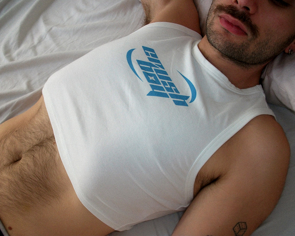 A man lying on a bed wearing a white cropped tank top with a blue 'crushboy' logo printed on the chest.