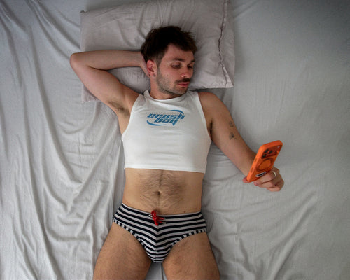 A man lying on a bed with white sheets, wearing a white cropped tank top with a blue graphic and black-and-white striped underwear, while holding an orange smartphone.