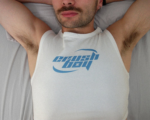 A man lying down wearing a white sleeveless tank top with a blue 'crushboy' graphic printed on the chest, set against a gray bedding background.
