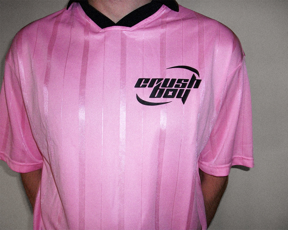 A pink short-sleeve athletic jersey with vertical stripes, a black collar, and a black 'crushboy' logo on the chest, worn by a person against a plain gray background.