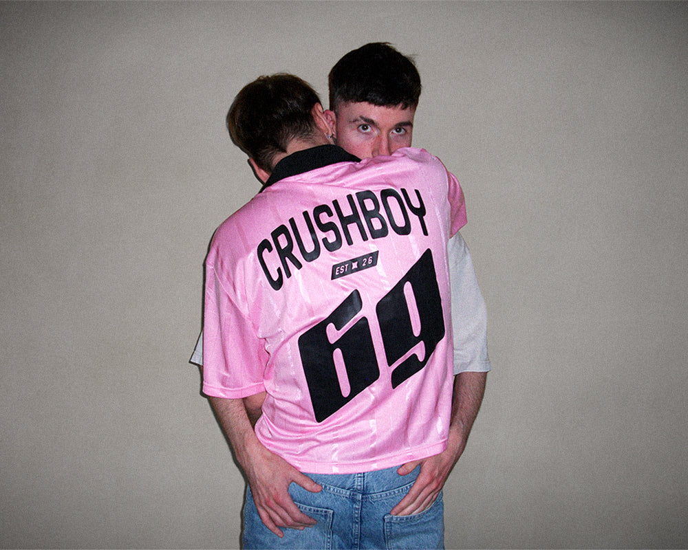 A man wearing a pink Crushboy jersey with the number 69 on the back, hugging another man against a plain beige wall.