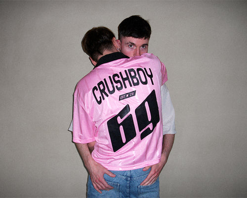 A man wearing a pink Crushboy jersey with the number 69 on the back, hugging another man against a plain beige wall.