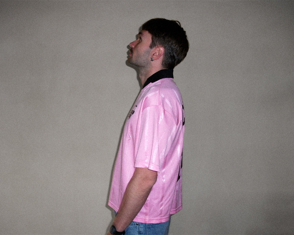 A man shown in profile wearing a light pink short-sleeve athletic jersey with a black collar, standing against a plain light gray wall.