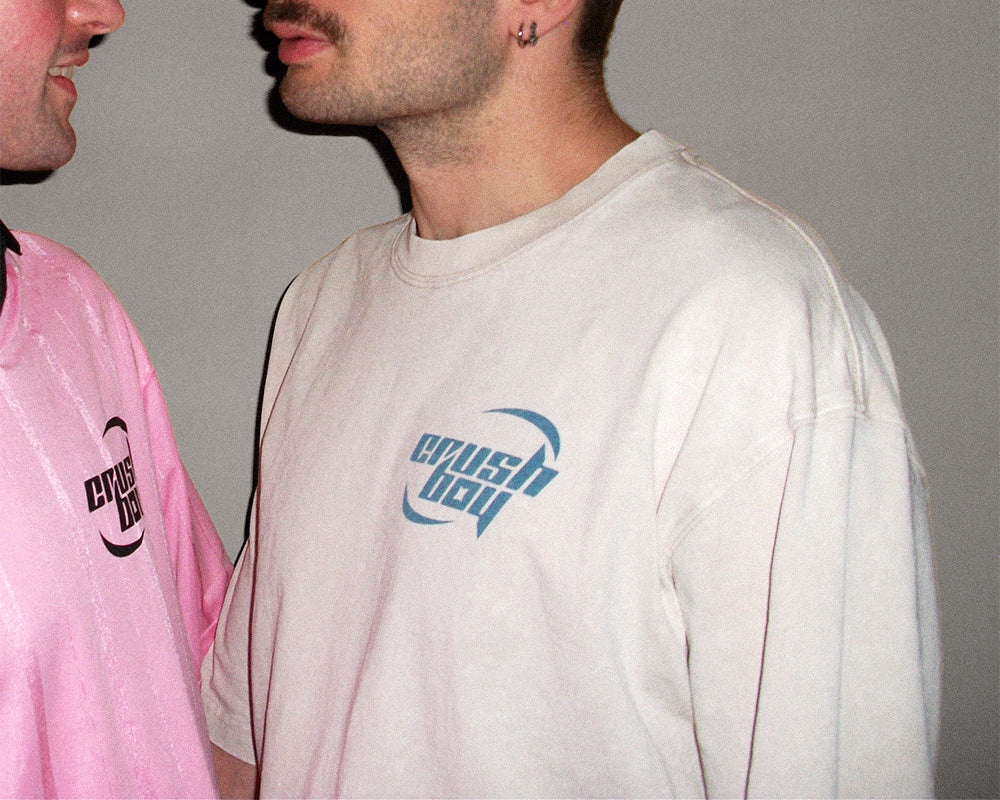 A man wearing a light beige crewneck sweatshirt with a blue 'crushboy' graphic on the chest, standing next to another person in a pink sweatshirt against a plain gray wall.