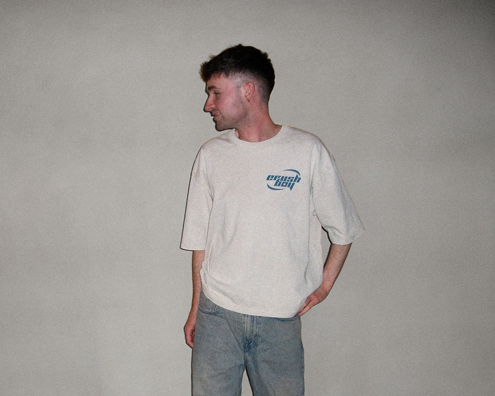 A man wearing an oversized off-white t-shirt with a blue 'crushboy' logo on the left chest, paired with light blue jeans, standing against a plain gray wall.