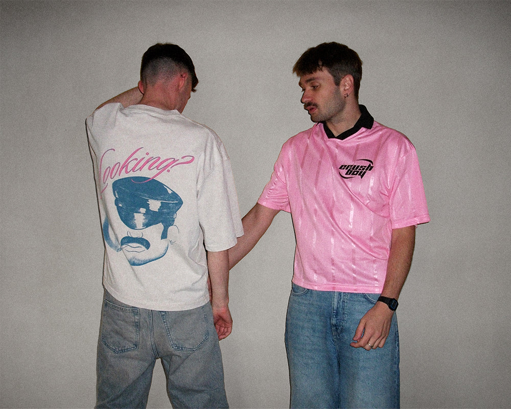 Two men standing against a plain light gray wall. The man on the left wears a white graphic T-shirt with a blue face print and pink script text, shown from the back.