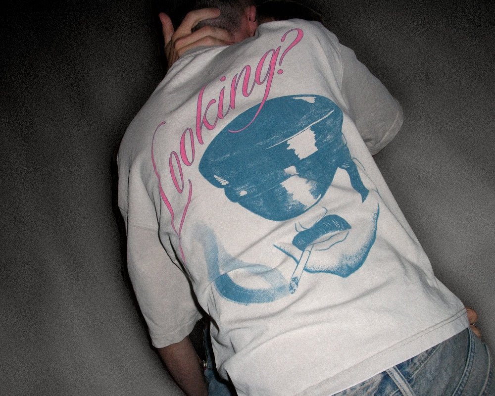 A person viewed from behind wearing a light gray t-shirt with a large graphic print on the back. The print features a blue stylized face wearing a hat and the word 'Looking?' in pink script.