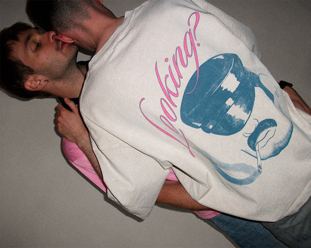 A man wearing a white graphic t-shirt with a blue and pink design, being kissed on the cheek by another man in a dimly lit indoor setting.
