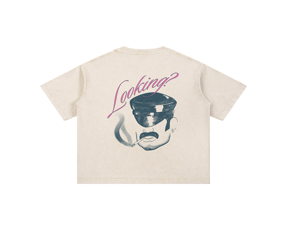 A cream-colored short-sleeve t-shirt with a graphic print on the back. The graphic features a stylized illustration of a face wearing a cap and smoking a cigarette, with the word 'Looking?' written in pink script above it.