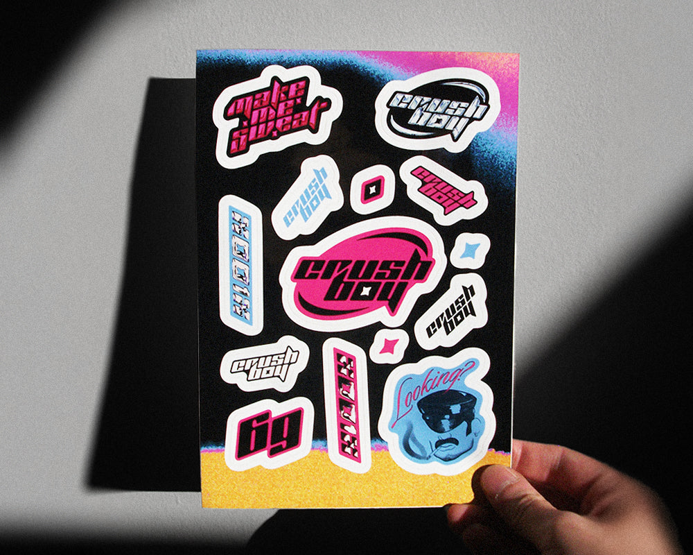 A hand holds a sticker sheet featuring various retro-style 'CRUSHBOY' and 'make me sweat' decals in pink, blue, white, and black, set against a gray background with dramatic shadows.