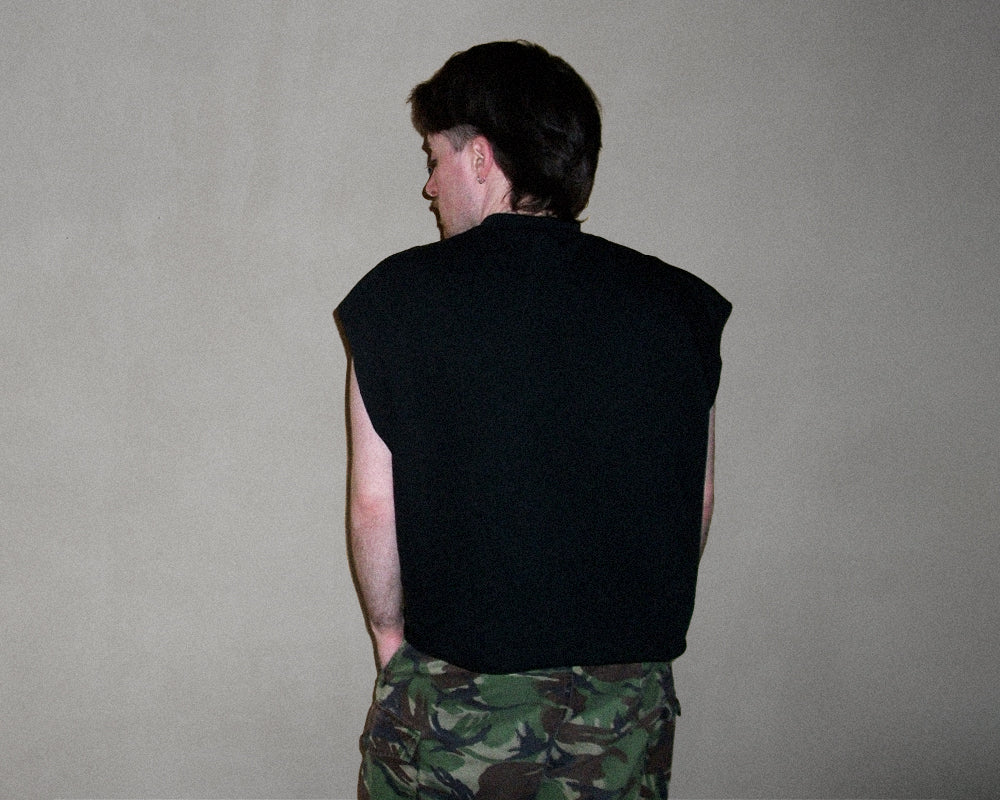 A man shown from the back wearing a black sleeveless top and green camouflage pants against a plain gray wall.