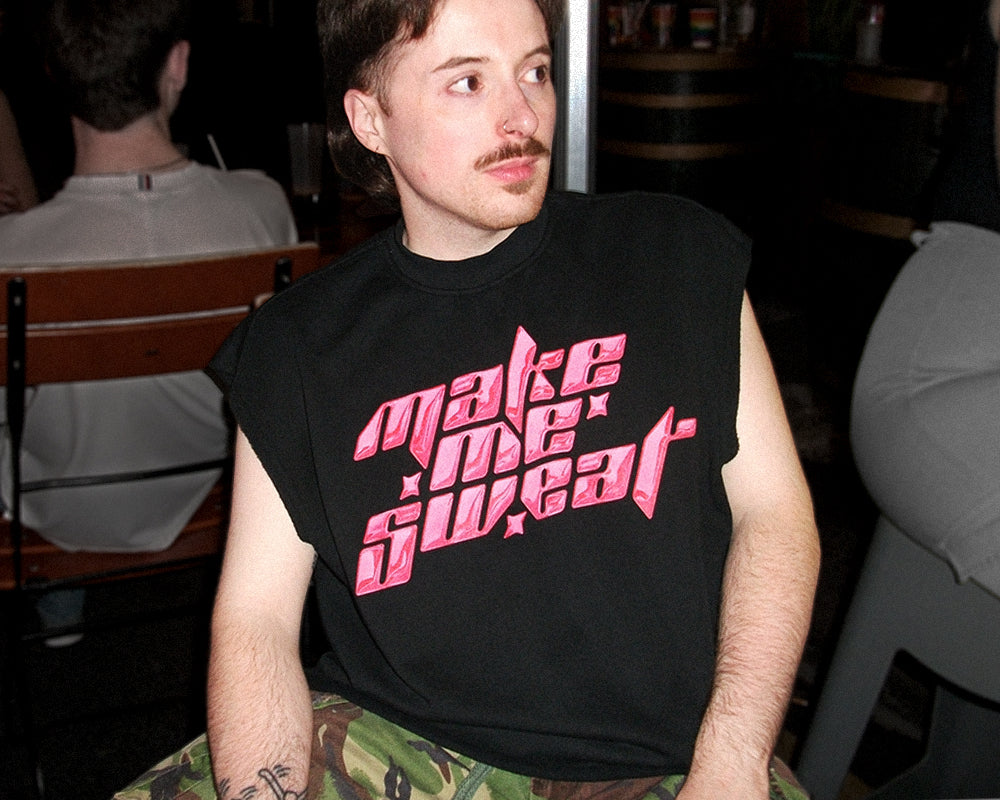 A man wearing a black sleeveless t-shirt with 'make me sweat' printed in pink stylized text, seated in a dimly lit smoking area.