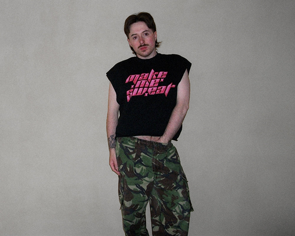 A man wearing a black sleeveless t-shirt with pink 'make me sweat' text and green camouflage cargo pants, standing against a plain gray wall.