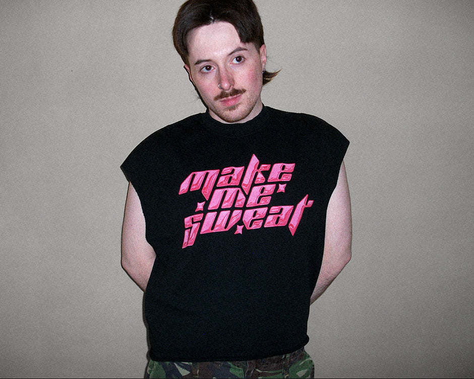 A man wearing a black sleeveless t-shirt with 'make me sweat' printed in pink metal-style lettering, standing against a plain gray wall.