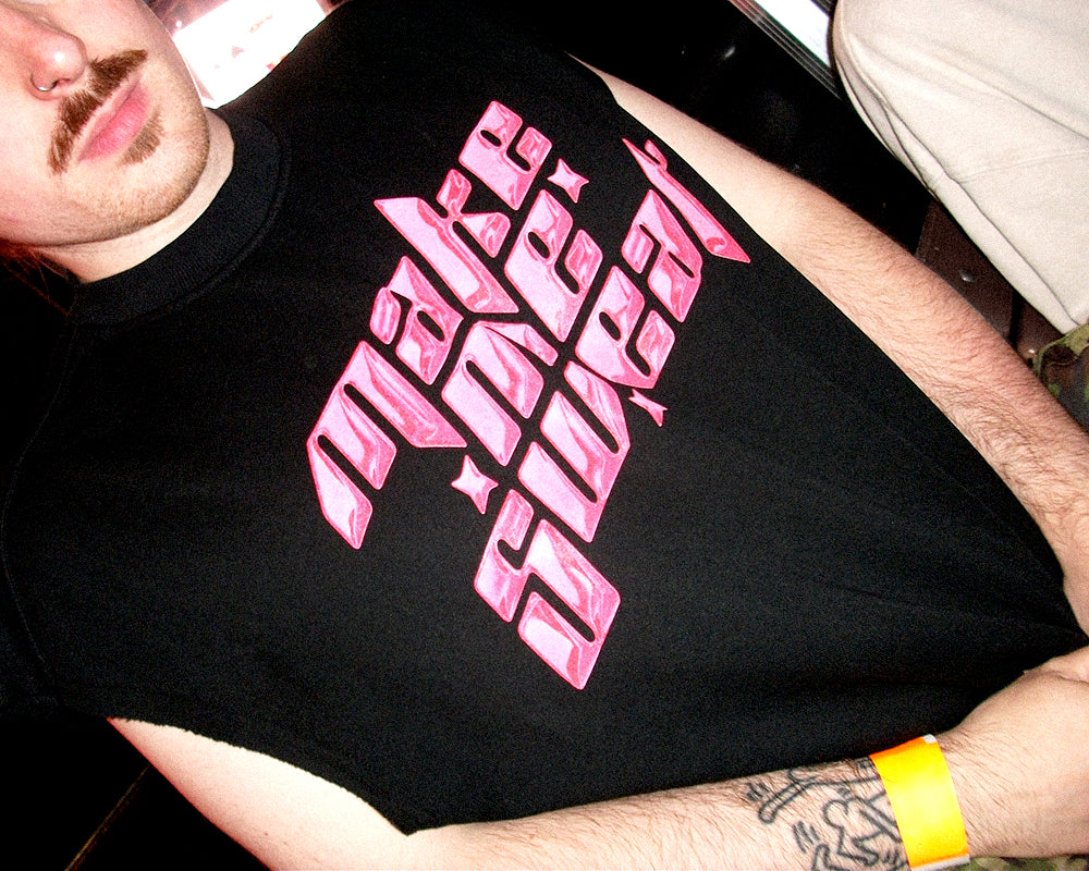 A close-up of a man wearing a black sleeveless t-shirt with a large pink metallic-style graphic text reading 'make me sweat' across the chest.