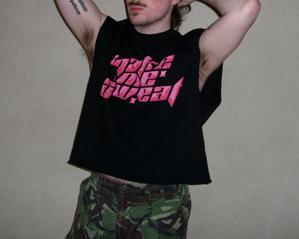 A man wearing a black sleeveless crop top with a pink graphic text design that reads 'make me sweat' in a stylized font, paired with green camouflage pants, against a plain beige wall.